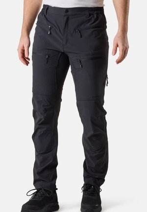 Homme portant un pantalon d'extérieur noir avec plusieurs poches zippées et des chaussures de sport noires, debout devant un fond uni.