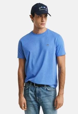 Helles blaues Baumwoll-T-Shirt mit Rundhalsausschnitt und kurzen Ärmeln. Getragen mit hellblauen Jeans, ausgestattet mit einem schwarzen Gürtel und einer marineblauen Mütze mit Logos.