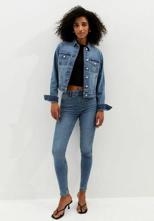 Femme aux cheveux bouclés portant une veste en denim bleu, un jean skinny taille haute, un top court noir et des sandales à talons noires, debout contre un mur blanc.