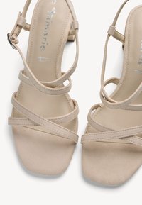 Sandales à talons hauts beiges avec un embout carré, dotées de lanières croisées et d'une boucle à la cheville. Le matériau a une texture douce, semblable au daim.