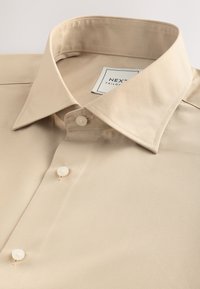 Béžová košile na míru s špičatým límečkem, texturovanou tkaninou a třemi bílými knoflíky. Značková etiketa viditelná uvnitř límce.