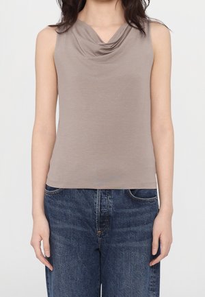 Frau trägt ärmelloses Taupe-Top mit Wasserfallausschnitt und blaue Jeans, steht mit entspannt hängenden Armen seitlich vor einfachem Hintergrund.