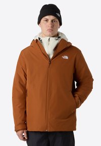 Chaqueta impermeable marrón con cierre de cremallera, capucha ajustable y forro de forro polar, combinada con un gorro de punto negro.
