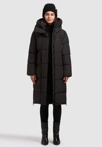 Sort pufferjakke med hætte, quiltet design, frontlynlås og trykknaplukninger. Kombineret med en matchende strikkemøsr og tætsiddende sorte bukser.