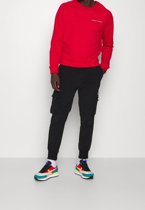 Muž v červené mikině, černých cargo kalhotách, bílých ponožkách a vícebarevných teniskách Puma stojí před jednobarevným bílým pozadím.