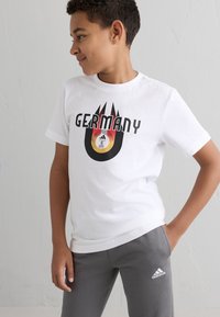 Camiseta de algodón blanca con "ALEMANIA" en letras negras en negrita, con un gráfico redondo colorido de un balón de fútbol y llamas debajo. Pantalones grises.