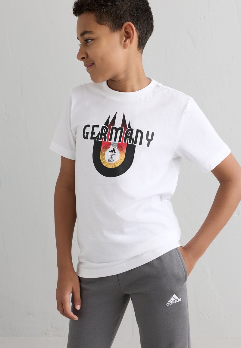 Camiseta de algodón blanca con "ALEMANIA" en letras negras en negrita, con un gráfico redondo colorido de un balón de fútbol y llamas debajo. Pantalones grises.