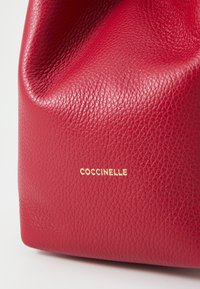 Raudona odinė rankinė su akmenukų tekstūra; turi išlenktą formą ir įspaustą auksinį "Coccinelle" logotipą.