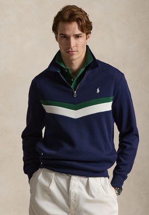Jeune homme portant un pull bleu marine à fermeture éclair quart avec des rayures en chevrons vertes et blanches, une chemise verte à col, un pantalon blanc et une montre-bracelet.