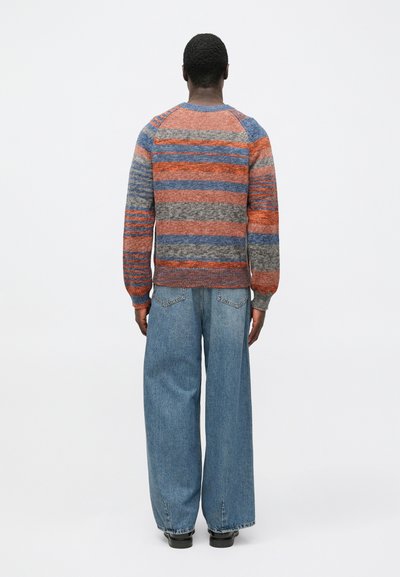 Pletený svetr s vícebarevnými pruhy v červené, modré, šedé a oranžové barvě. Žebrované manžety a lem, doplněné volnými modrými džínami a černou obuví.