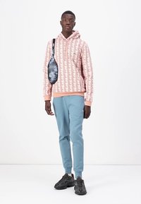 Felpa con cappuccio rosa chiaro con un motivo geometrico, abbinata a pantaloni della tuta blu chiaro e scarpe sportive nere. Borsa a tracolla blu e grigia.