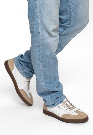 Beine in hellblauen Jeans und weißen Turnschuhen mit beigen Wildlederakzenten und braunen Sohlen vor weißem Hintergrund.