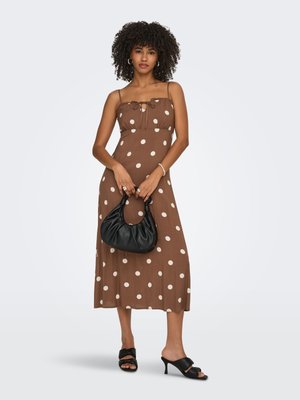 Vrouw met krullend haar die een bruine midi-jurk met stippen draagt, een zwarte handtas vasthoudt en zwarte sandalen met hakken draagt tegen een effen achtergrond.