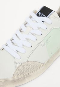Sneaker bianca in pelle con accenti verde menta, punta beige testurizzata, lacci bianchi piatti e etichetta nera con logo sulla linguetta.