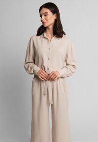 Beżowa lniana koszula z długimi rękawami, kołnierzykiem i złotymi guzikami. Połączona z spodniami o wysokim stanem i szerokich nogawkach, związanymi w talii.