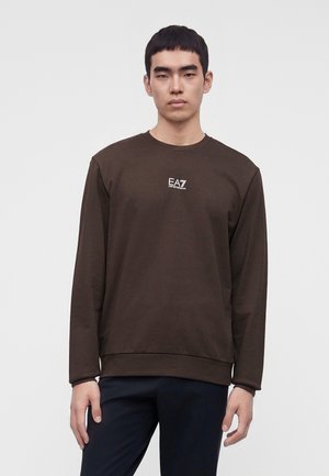 Mladý muž s krátkymi čiernymi vlasmi nosí tmavohnedú košeľu s dlhým rukávom s bielym logom EA7 Emporio Armani na hrudi, stojí na svetlom pozadí.
