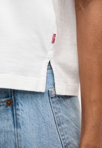 Ourlet d'un t-shirt en coton blanc avec une petite étiquette rouge Levi's, fente latérale, porté sur la poche et le bouton d'un jean en denim bleu clair.