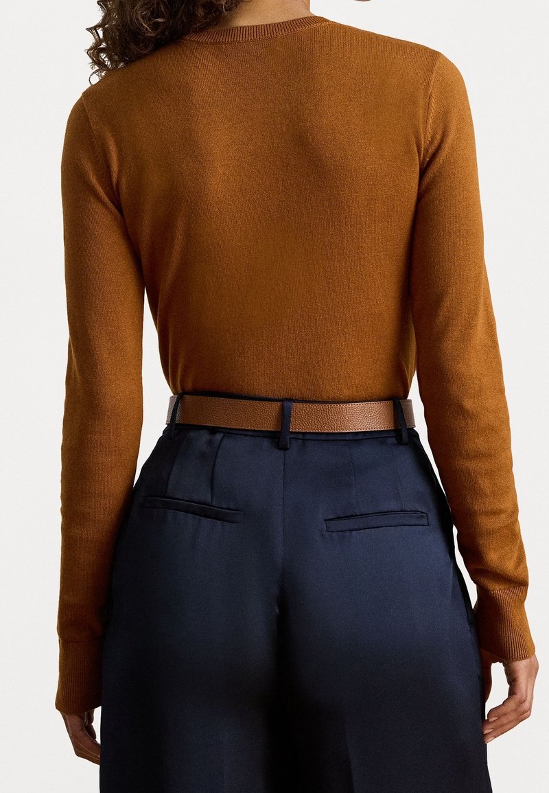 Personne portant un pull marron à manches longues rentré dans un pantalon bleu foncé taille haute avec une ceinture marron, vue de dos.