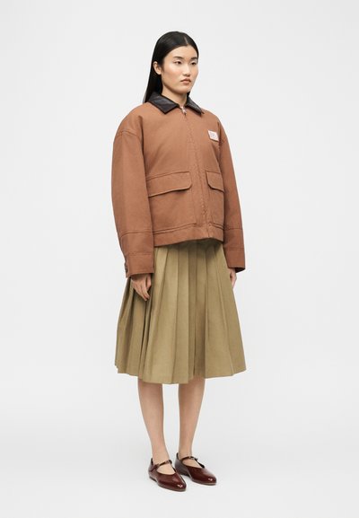 Γυναίκα που φορά καφέ μπουφάν με μαύρο γιακά, φούστα λαδί με πιέτες και σκούρα καφέ παπούτσια Mary Jane, στέκεται μπροστά σε ένα απλό φόντο.