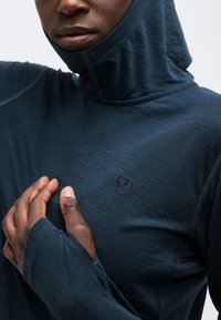 Close-up of a person wearing a navy hooded top with thumb holes, hand placed on chest, showing textured fabric and a small embroidered logo.