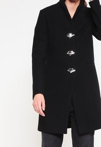 Kvinna klädd i en svart kappa med hög krage och tre stora silverkläppen som fäster framtill, står mot en neutral bakgrund.