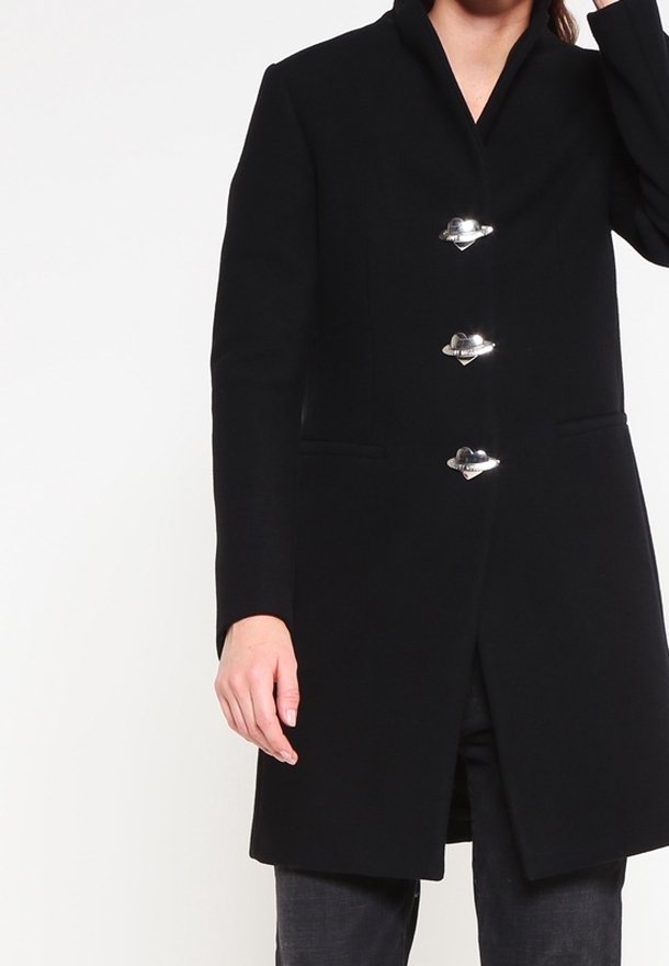 Kvinna klädd i en svart kappa med hög krage och tre stora silverkläppen som fäster framtill, står mot en neutral bakgrund.
