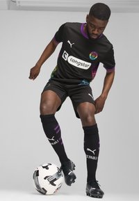 Equipación de fútbol negra con patrones multicolores en las mangas, presenta un logo blanco en el pecho, pantalones cortos negros y calcetas negras con rayas moradas.