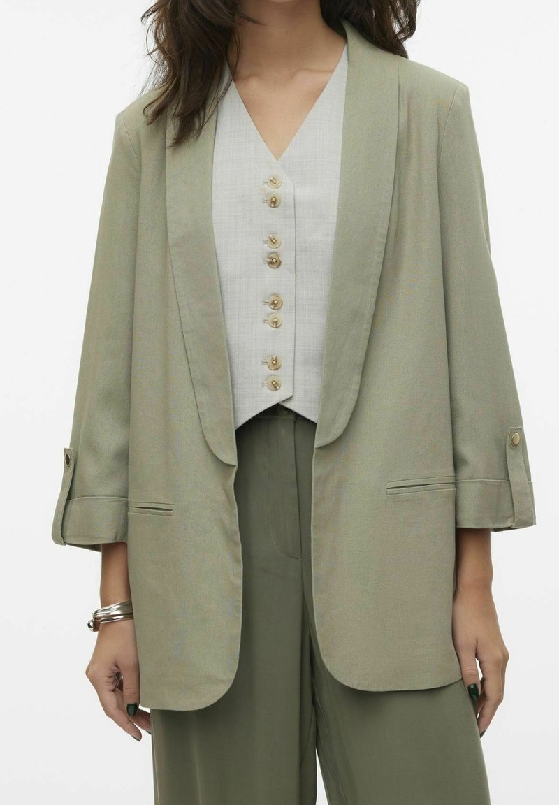 Femme portant un blazer vert clair avec manches retroussées sur un gilet blanc boutonné et un pantalon vert assorti, debout, les bras détendus.