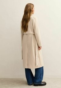 Abrigo largo beige hecho de una tela suave con cintura ceñida por un cinturón, que presenta una silueta recta y costuras mínimas. Combinado con jeans de pierna ancha azules.