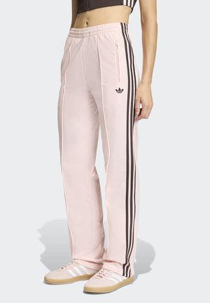 Persona vistiendo pantalones deportivos Adidas color rosa claro con franjas negras laterales, un bolsillo lateral con cremallera y zapatillas a juego rosas con rayas blancas.