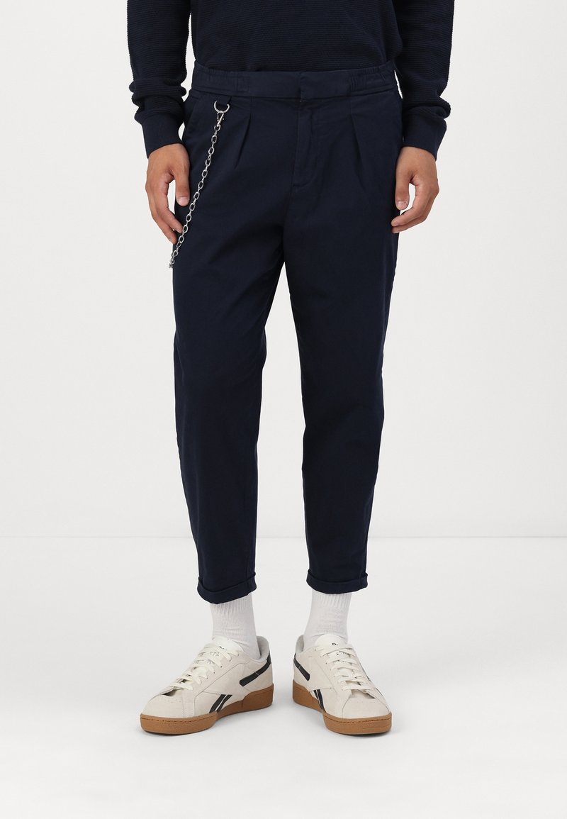 Námořnicky modré chino kalhoty s záhyby, zdobené kovovým řetízkem na pase. Kombinované s krémovými teniskami a bílými ponožkami.