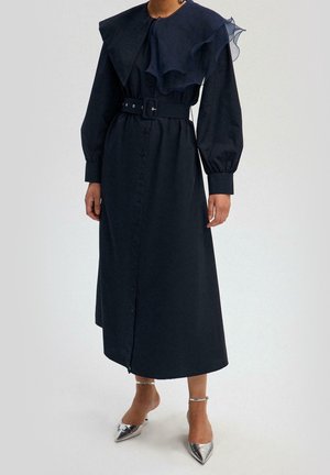 Donna che indossa un abito midi nero a manica lunga con cintura, ampio colletto e dettagli in tessuto trasparente arricciato, abbinato a tacchi a punta argentati.