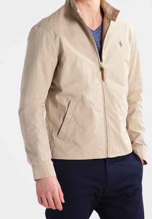 Beige Jacke mit Reißverschluss und Stehkragen, getragen über einem blauen V-Ausschnitt-Shirt, kombiniert mit dunkelblauen Hosen.