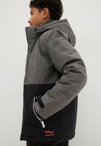 Veste zippée grise et noire avec un tissu texturé, une capuche, des poches latérales et un patch logo sur le côté inférieur.