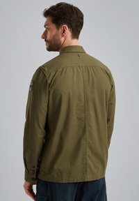 Camicia verde oliva con bottoni, in tessuto leggero, maniche lunghe, design con yoke posteriore, dotata di polsini con bottone e una toppa con logo sulla spalla.