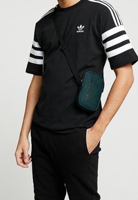 Černé bavlněné tričko s bílými pruhy, s logem Adidas. Použitá je malá zelená crossbody taška s texturovaným brandingem.