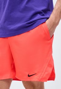 Άνδρας με φωτεινό πορτοκαλί σορτς Nike, με το χέρι στην τσέπη και μοβ πουκάμισο, τονίζοντας το πλάι και το λογότυπο του σορτς.