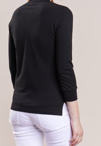 Svart långärmad tröja med hög halsringning, slät textur och detalj med delad fåll, i kombination med vita åtsittande jeans.