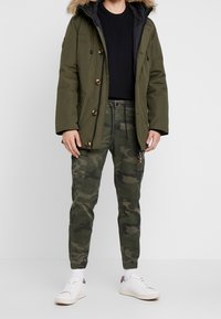 Parka verde con capucha forrada de piel, interior negro, botones dorados; combinada con pantalones cargo de camuflaje y zapatillas blancas. Atuendo casual y práctico.