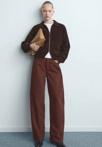 Chaqueta marrón con cremallera y textura sobre una camisa blanca, combinada con pantalones burdeos de talle alto y pierna ancha. Sosteniendo un bolso de mano color marrón claro.