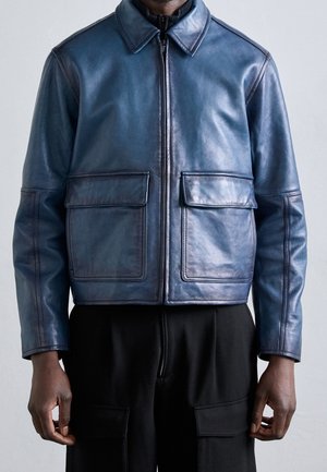 Homme portant une veste zippée en cuir bleu avec de grandes poches à rabat sur un pantalon noir avec des poches latérales.
