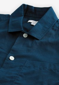 Camisa azul marino con cuello clásico, dos botones blancos y un bolsillo en el pecho. Fabricada con un tejido texturizado. Etiqueta visible en el interior del cuello.