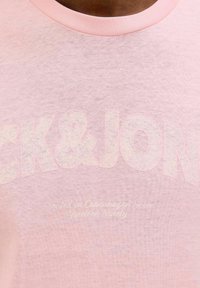Lichtroze T-shirt met een textuurafwerking. Heeft vetgedrukte witte letters "CK&JN" en kleinere tekst "Gemaakt in Kopenhagen." Eenvoudige ronde halslijn.