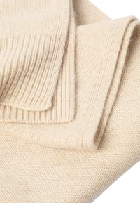 Béžová pletená látka s mäkkou textúrou, na okrajoch s rebrovaným vzorom. Záhyby vytvárajú hĺbku a rozmer.