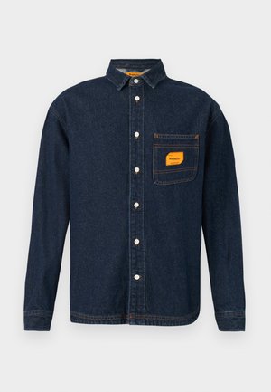 Donkerblauwe denim shirt met button-down kraag, oranje label op de borstzak en witte knopen. Zachte textuur met contrasterende stikseldetails.