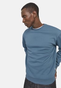 Sweat-shirt bleu clair en tissu de coton doux, avec un col rond et des poignets côtelés, associé à un jean en denim foncé. Design simple, sans motifs.