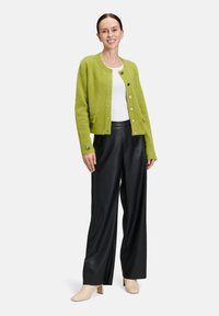 Femme souriante, portant un cardigan vert citron à boutons, un haut blanc, un pantalon large noir et des bottes à talons beiges, se tenant devant un fond blanc.