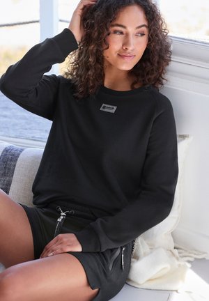 Young woman with curly hair wearing a black sweatshirt and shorts sitting by a window with pillows and a light blanket behind her.