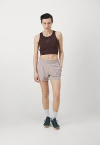 Rudas trumpas viršus su logotipu, šviesiai smėlio spalvos šortai su elastinga juosmens dalimi, baltos kulkšnies kojinaitės ir tamsiai žalios sportbačiai. Paprastas fonas.