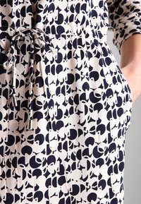 Puuvillamekko, jossa on kerma beige tausta ja rohkea tummansininen abstrakti kuvio. Mekossa on narutettava vyötärö, sivutaskut ja kolmannelta neljännekseltä hihat.